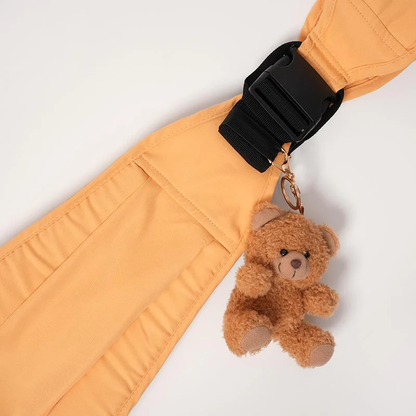 Doudou ours marron avec mousqueton accroché sur un porte-bébé orange, accessoire de portage doux et rassurant pour bébé