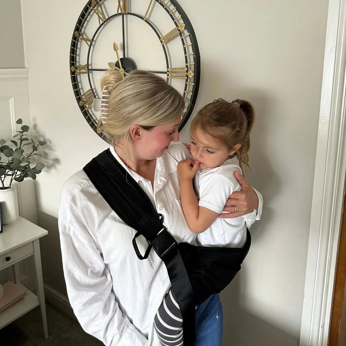 Maman portant son enfant dans le porte-bébé HelloBiboo à la maison, enfant rassuré et confortablement installé contre elle.